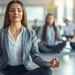 Junge Frau in Business-Kleidung sitzt in Yoga-Pose auf dem Fußboden, hinter ihr verschwommen sieht man noch mehr Frauen