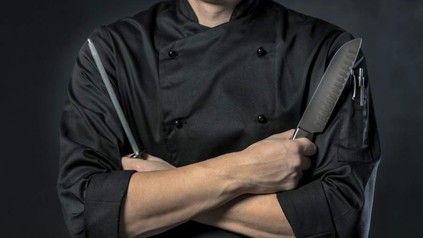 Ein schwarz gekleideter Koch mit Messer und Messerschärfer verschränkt die Arme