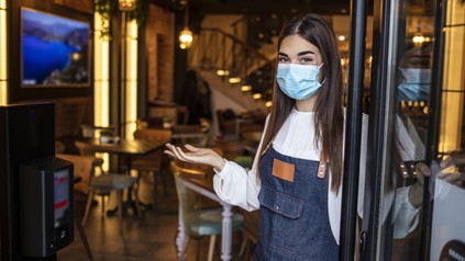 Portrait of a young Caucasian female waitress wearing an apron, face mask and gloves, standing at the entrance of a drink establishment and welcoming customers of the store.