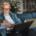 Rüstiger Rentner sitzt mit Laptop und Notizbuch auf der Couch