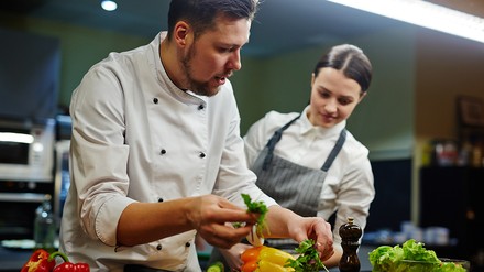 EIn Koch arbeitet mit Gemüse, eine Küchenhilfe schaut ihm über die Schulter