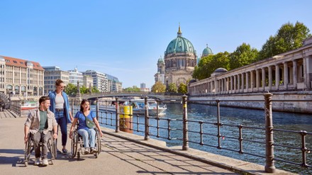 Drei Menschen, darunter zwei Rollstuhlfahrer, spazieren gemeinsam am Berliner Spreeufer mit Blick auf den Berliner Dom und die Museumsinsel
