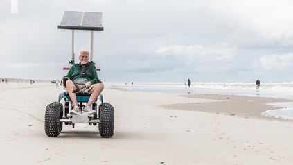 Mann fährt mit einem geländegängigen, solarbetriebenen Strandrollstuhl über den Strand auf Juist; im Hintergrund Nordsee und Spaziergänger.