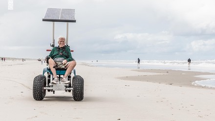 Mann fährt mit einem geländegängigen, solarbetriebenen Strandrollstuhl über den Strand auf Juist; im Hintergrund Nordsee und Spaziergänger.
