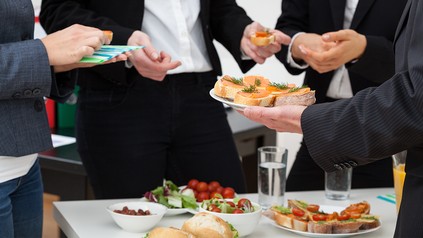Mehrere Personen in Businesskleidung greifen zu belegten Broten und Fingerfood an einem Buffet bei einem Büro-Event.