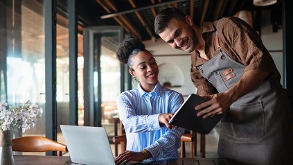 Restaurantmitarbeiter zeigt einer Kollegin am Tisch auf einem Tablet Zahlen, während ein Laptop geöffnet ist.