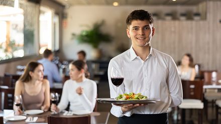 Junger Kellner im Restaurant präsentiert Salat und Wein auf einem Tablett – Symbolbild für junge Beschäftigte im Gastgewerbe und die Generation Z.
