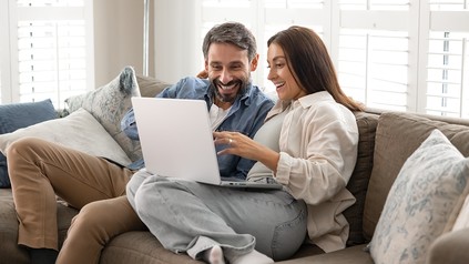 Paar sitzt entspannt auf einem Sofa und nutzt gemeinsam einen Laptop, um online eine Reise zu planen.