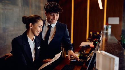 Zwei gut gelaunte Hotelmitarbeiter arbeiten gemeinsam am Empfang eines Hotels und schauen auf einen Computerbildschirm