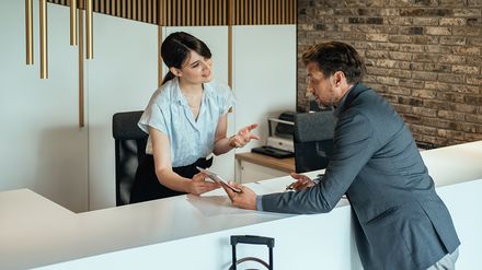 Hotelmitarbeiterin zeigt einem Gast an der Rezeption digitale Informationen auf einem Tablet