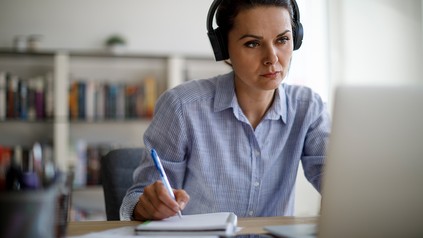 Frau mit Kopfhörer vor dem Laptop, schreibt Notizen auf