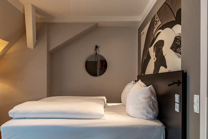 Hotelzimmer mit Doppelbett, weißen Bettwaren, rundem Wandspiegel und großformatigem Wandbild.