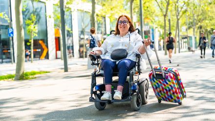 Frau im Elektrorollstuhl zieht einen Rollkoffer auf einer barrierefrei gestalteten Straße mit Passanten im Hintergrund.