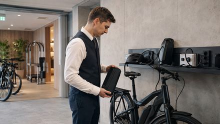 Hotelmitarbeiter prüft einen E-Bike-Akku an einer Ladestation in einem modernen Fahrradraum.