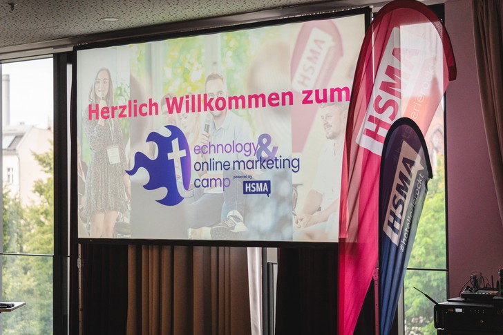 Willkommensprojektion des Technology & Online Marketing Camps neben zwei Fahnen der HSMA.