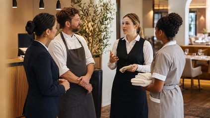 Vier Mitarbeiter aus Rezeption, Küche, Service und Housekeeping stimmen sich in einem Hotelrestaurant vor der Schicht ab.