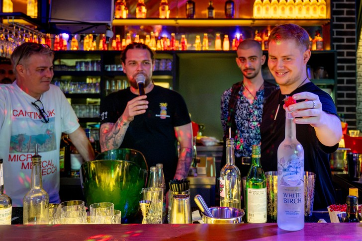 Vier Männer stehen hinter einer Bar mit Flaschen und Gläsern; einer hält ein Mikrofon, ein anderer zeigt einen Drink.