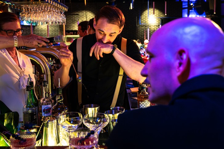 Ein Mann mit Hosenträgern steht hinter der Bar und wird von einer Frau mit Mikrofon begleitet.