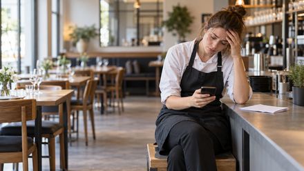 Ein Mitarbeiter im Restaurant sitzt erschöpft an der Theke und informiert den Arbeitgeber per Smartphone über seinen Ausfall.