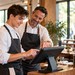 Ein junger Restaurantmitarbeiter lernt mit Unterstützung eines erfahrenen Kollegen die Bedienung eines digitalen Kassensystems.