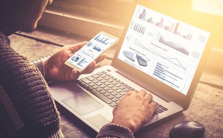 Mann sitzt vor Laptop und Handy und vergleicht Diagramme