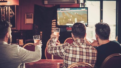 Drei junge Männer mit Bier in der Hand bejubeln ein Fußballspiel im Fernseher