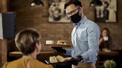 Freundlicher Kellner mit Mund-Nasen-Maske bedient eine Frau im Restaurant