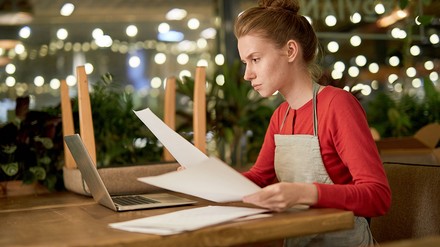Junge Gastronomin sitzt vor einen geöffneten Laptop und hält Papierdokumente in den Händen