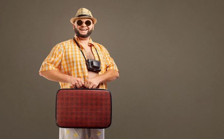 Ein dicker Mann mit offenem Hemd, Kamera um den Hals und altmodischem Koffer