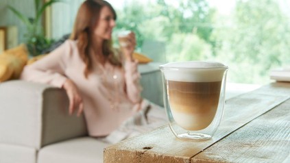 Ein modernes Glas mit Kaffee gefüllt und eine Dame im Hintergrund, die gerade aus einem solchen Glas trinkt