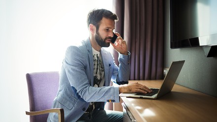Businessmann sitz im Hotelzimmer an einem Tisch, telefoniert und tippt etwas in seinen Laptop
