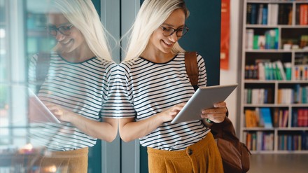 Junges Mädchen steht lächelnd mit einem Tablet in der der Bibliothek