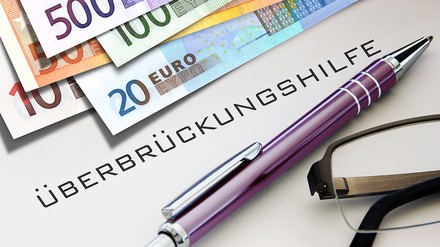 Euro-Geldscheine, ein violetter Kugelschreiber, eine Lesebrille und das Wort „Überbrückungshilfe“