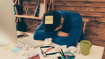 Müder junger Mann hat die Arme erschöpft auf den Schreibtisch gelegt und seinen Kopf darauf gestützt, auf seinem Kopf klebt ein gelbes Post-it mit der Aufschrift Help