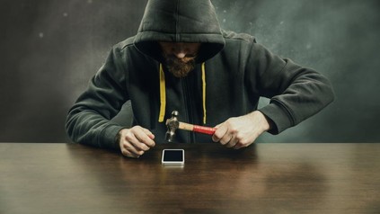 Mann mit Kapuzenpullover schlägt mit Hammer auf ein Smartphone