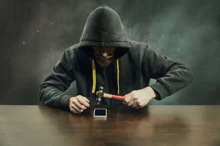 Mann mit Kapuzenpullover schlägt mit Hammer auf ein Smartphone