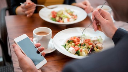 Zwei Personen essen gemeinsam während eine der beiden das Smartphone in der Hand hält
