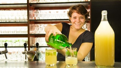Eine lächelnde Barmitarbeiterin gießt ein Getränk aus einer großen Flasche in ein Glas