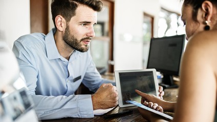 Ein Hotelmitarbeiter an der Rezeption zeigt einer jungen Frau etwas auf dem Tablet