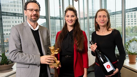 Zwei Frauen und ein Mann stehen vor einer Fensterfront und halten eine große Weinflasche und den Goldenen Bär, den Hauptpreis der Berlinale, in der Hand