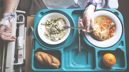 Blick auf ein Essenstablett mit mehreren Tellern, von denen eine junge Patientin im Krankenhaus isst