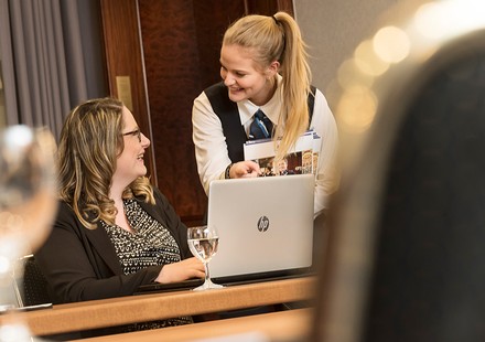 Eine Hotelmitarbeiterin spricht lächelnd mit einer Besucherin an der Rezeption vor einem Laptop