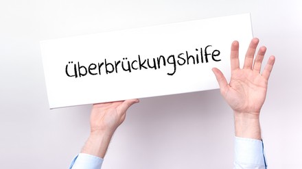 Ein untergehender Unternehmer hält ein Schild hoch, auf dem „Überbrückungshilfe“ steht