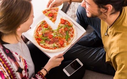Ein junges Pärchen isst eine Pizza neben der ein Smartphone liegt