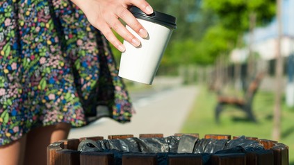 Nahaufnahme einer Frauenhand, die einen Einweg-Kaffeebecher in einen Mülleimer im Park wirft