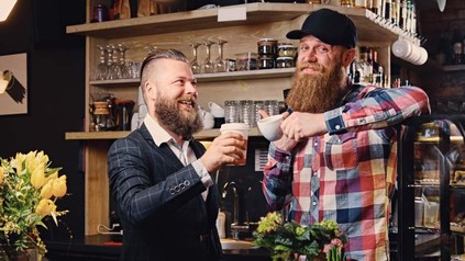 Zwei bärtige Männer, die sich hinter einer Restauranttheke mit Kaffeetassen zuprosten