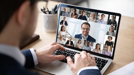 Man sitzt vor Laptop auf dem eine Videokonferenz zu sehen ist