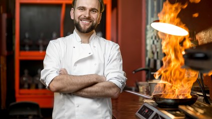 Koch steht zufrieden mit verschränkten Armen in der Küche, während im Hintergrund eine Pfanne auf dem Herd brennt