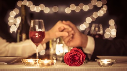 Ein Mann und eine Frau sitzen am Valentinstag an einem romantisch gedeckten Tisch im Restaurant und halten sich an der Hand, eine Rose liegt auf dem Tisch