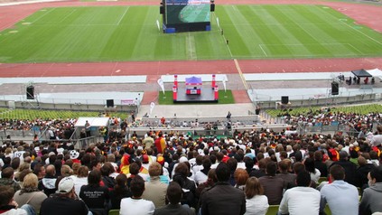 Ein gut besuchtes Fußballstadion auf dessen Rasen eine große Leinwand für Public Viewing aufgebaut ist